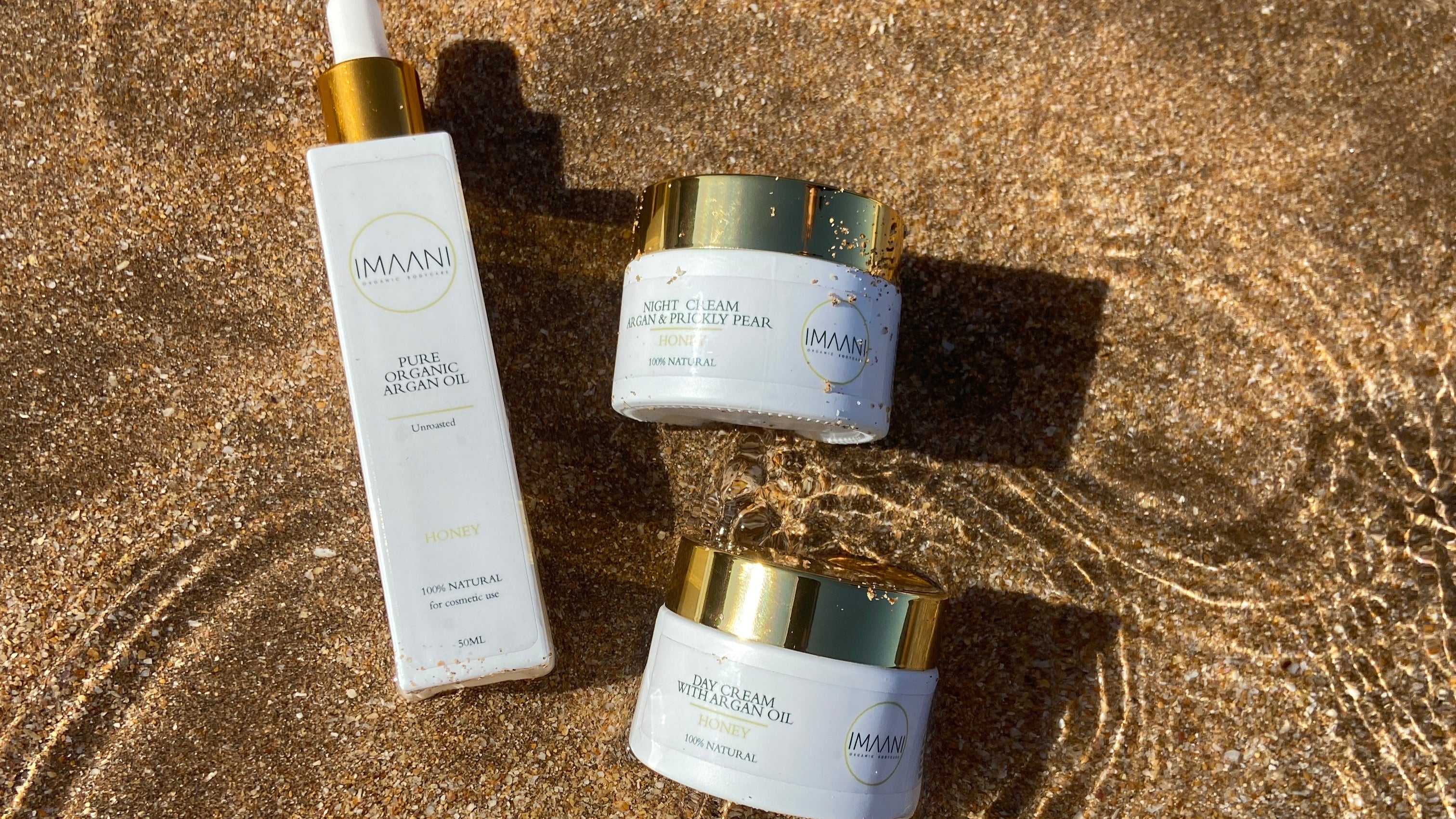 IMAANI pure argan oil bottle, Day and Night Cream on a sandy beach with ripples of water.